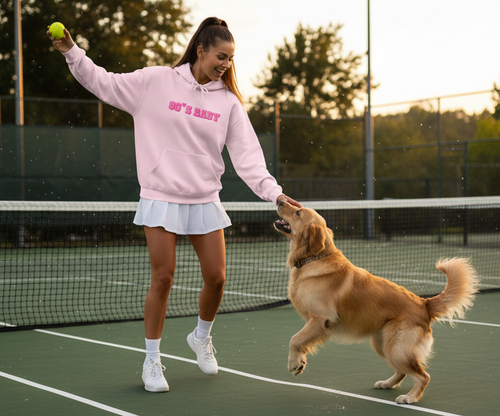 Woman in pink hoodie and white skirt playing with a dog on a tennis court