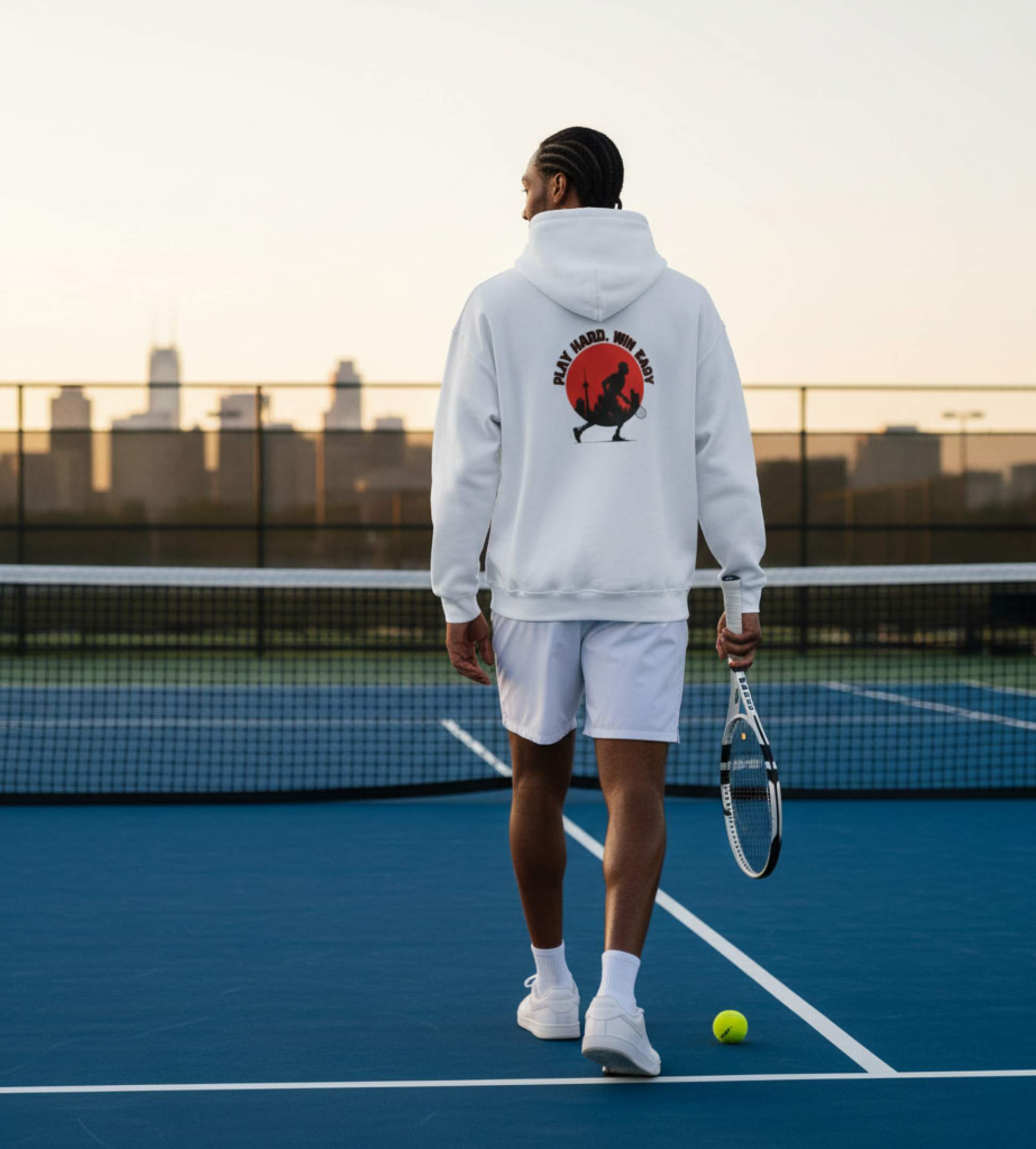 Person on a tennis court wearing a hoodie with a graphic design, holding a tennis racket, and a ball.