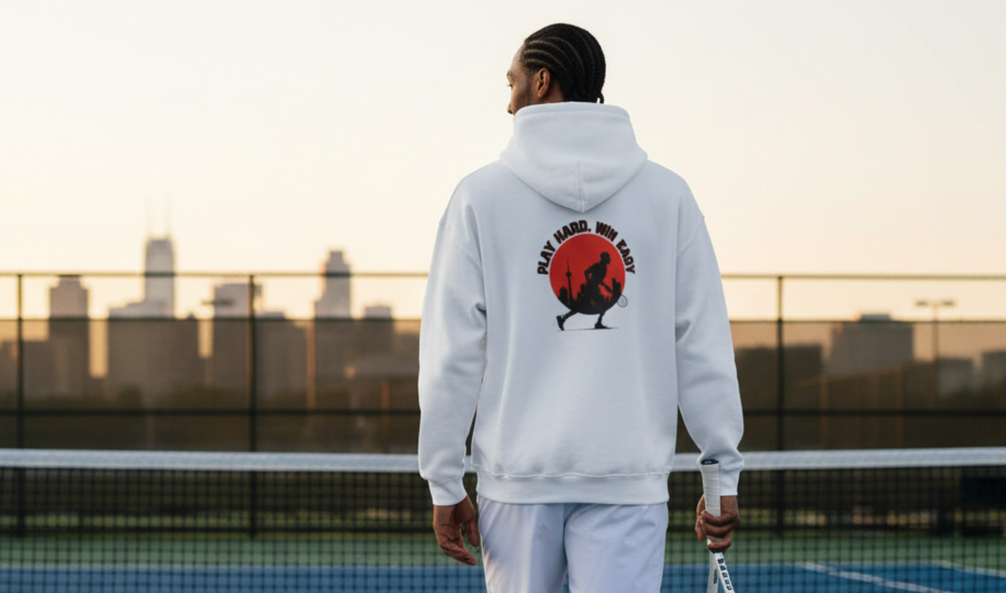 Person on a tennis court wearing a hoodie with a graphic design, holding a tennis racket, and a ball.