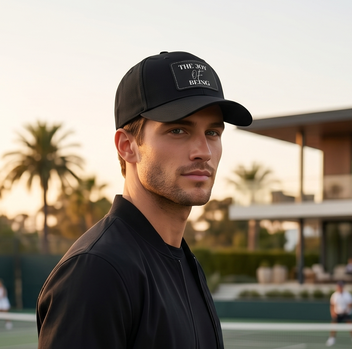 Man in black jacket and cap standing on a tennis court with palm trees and building in the background